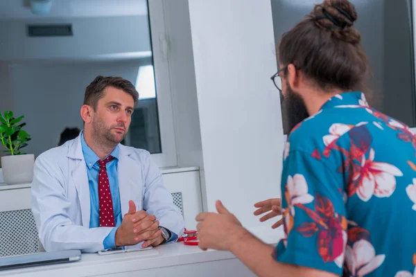 Un médico está hablando con un paciente hipster en el consultorio y le explica —  Fotos de Stock