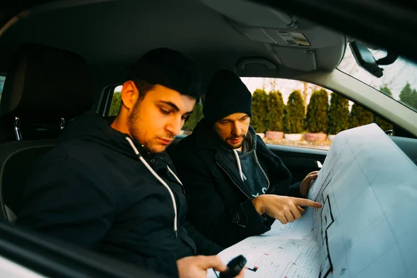 Zwei bewaffnete Banditen, die in einem Auto sitzen und ihren nächsten Raub planen, während sie auf eine Stoppuhr zählen, wie viel Zeit ihnen für den Raub bleibt, ihre Waffen zeigen und auf den Bauplan zeigen. — Stockbild Zwei bewaffnete Banditen sitzen in einem Auto und planen ihren nächsten Raubüberfall, — Stockfoto