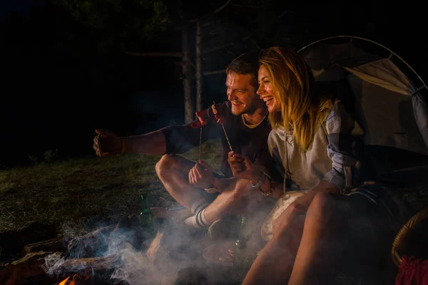 Genç çift kamp ateşinde sosis pişiriyor ve bira içiyor. — Stok fotoğraf