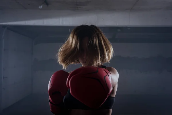 Silhouette portrait of young attractive fitness woman exercising punching with red boxing gloves in a garage