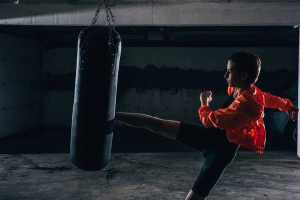 年轻的轮廓女性在车库里用拳击袋练习高踢腿 — 图库照片
