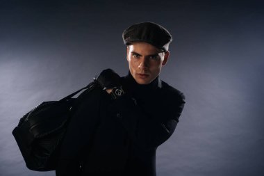 Portrait of a young man with sharp jawline wearing a newsboy cap, holding a man bag while posing in studio on a white background. clipart