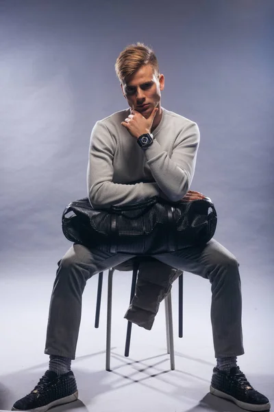 Informally ( casual ) dressed blonde model wearing a white sweater and holding a black man bag is posing on a white background while sitting on a chair.