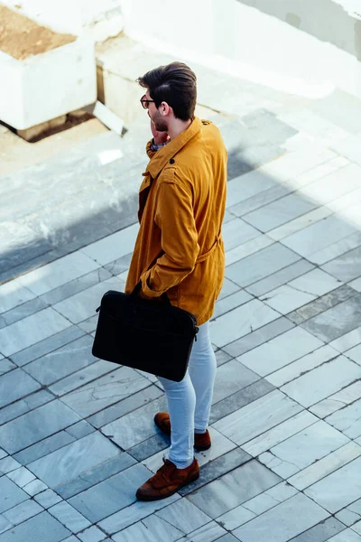 Urban businessman in yellow coat holding briefcase while walking down in the city