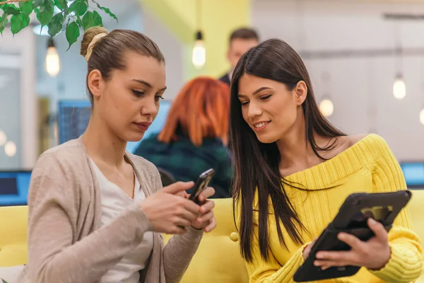 Multiethnic colleagues blond caucasian and Latina dark haired Handsome businesswomen (employee) sitting on a yellow couch while working on a laptop in a modern startup company