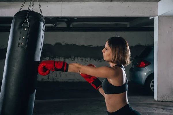 ガレージの中にボクシングバッグをパンチ強い若い女性のプロフィールビュー — ストック写真