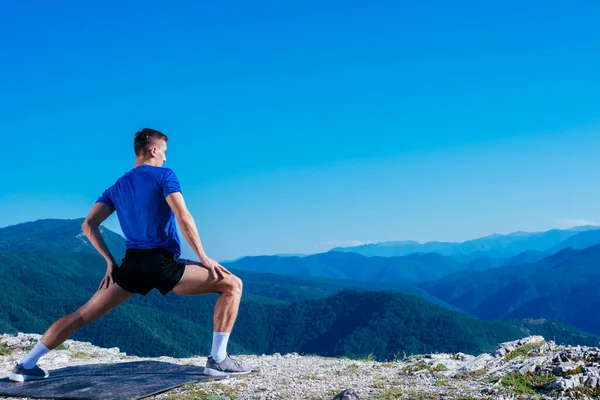 穿着蓝色干合身衬衫的金发运动员站在悬崖顶上伸展身体 准备锻炼 那里的景色令人叹为观止 — 图库照片