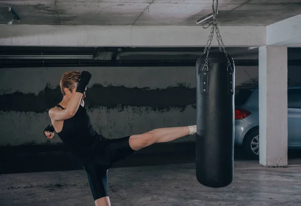 スポーツウェアを着た白人女性と黒の包帯がガレージの中でボクシングバッグを蹴って — ストック写真
