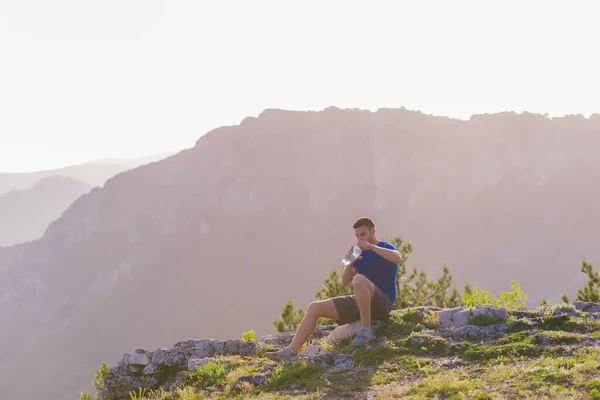 英俊的运动员穿着蓝色的衬衫和灰色的短裤 坐在石峰上 凝视着令人惊叹的山线和美丽的湖水 — 图库照片