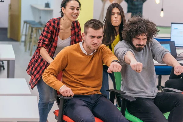 Team building and office fun.Four young cheerful businesspeople in smart casual wear having fun while racing on office chairs and smiling.