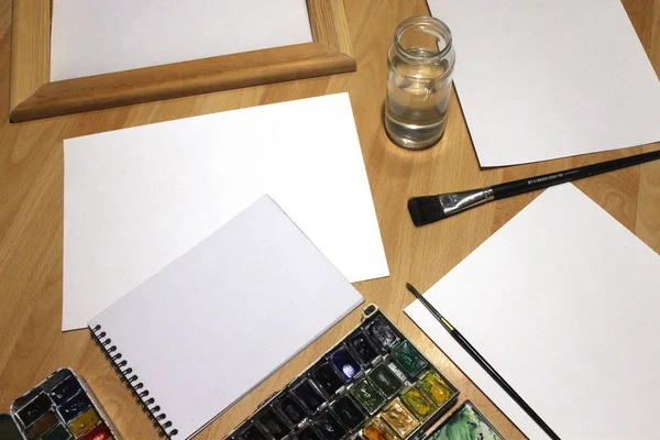 Notebook, modell, grafika, akvarell festék, paletta, pohár vizet és egy ecseteket fel. Művészi munkaeszközök a fehér fa asztal. — Stock Fotó