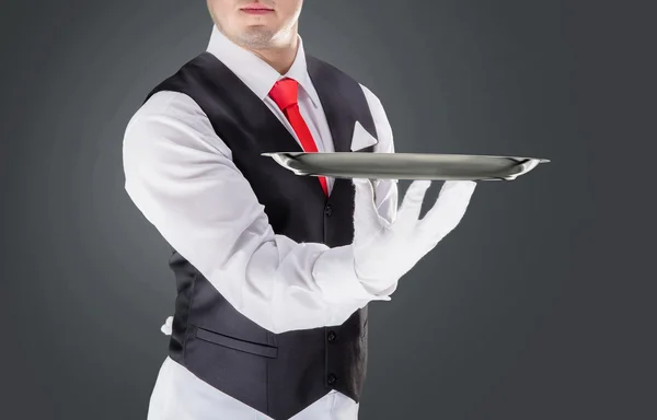 Handsome young waiter in gloves and red cravat holding empty tray — ストック写真