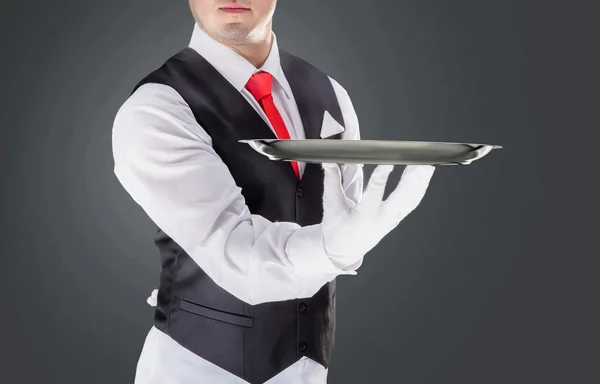 Bonito jovem garçom em luvas e vermelho cravat segurando bandeja vazia — Fotografia de Stock