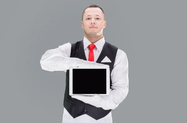 Bonito jovem garçom em luvas e vermelho cravat segurando comprimido — Fotografia de Stock