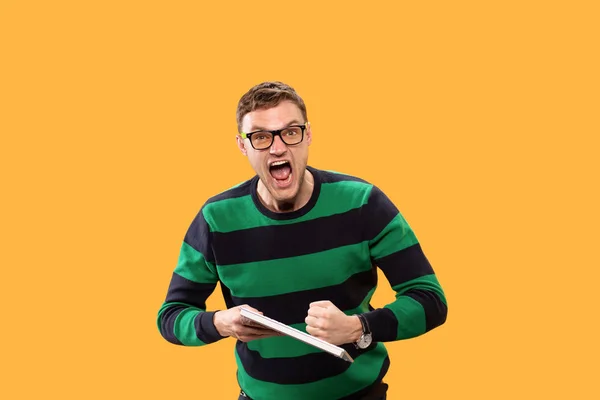 Angry young man holding a keyboard in his hands and trying to break it on yellow background. He screams heavily and looks at the camera. Studio photo. — 스톡 사진