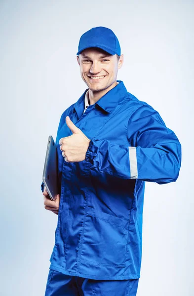 Ritratto di giovane lavoratore in uniforme blu. Tenendo in mano un portatile e mostrando il pollice in alto. Movimento a posto. Isolato su sfondo grigio con spazio di copia. Espressione del volto umano, emozione. Concetto aziendale . — Foto Stock