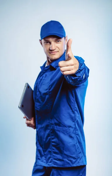 Porträtt av en ung arbetare i blå uniform. Hålla en laptop i handen och peka finger åt dig tittar på kameran. Isolerad på grå bakgrund med kopieringsutrymme. Mänskligt ansiktsuttryck, känslor. Affärsidé. — Stockfoto