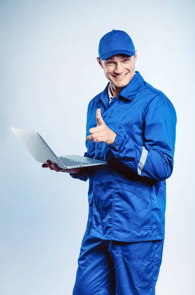 Porträtt av en ung arbetare i blå uniform. Hålla en laptop i handen och peka finger åt dig tittar på kameran. Isolerad på grå bakgrund med kopieringsutrymme. Mänskligt ansiktsuttryck, känslor. Affärsidé. — Stockfoto