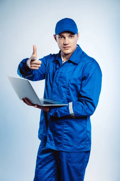 Retrato de un joven trabajador con uniforme azul. Sujetando un portátil en su mano y señalándote con el dedo mirando a la cámara. Aislado sobre fondo gris con espacio de copia. Expresión de rostro humano, emoción. Concepto empresarial . — Foto de Stock