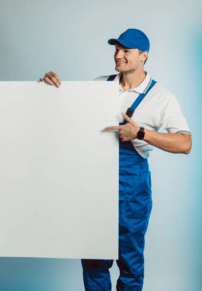 Imagem de um jovem trabalhador sorrindo segurando banner branco vazio e aponta para ele . — Fotografia de Stock