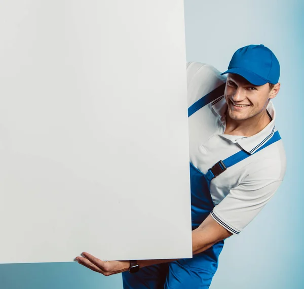 Imagen burlona de un joven trabajador sonriente sosteniendo una pancarta blanca vacía. Fondo blanco o azul. Concepto empresarial . — Foto de Stock