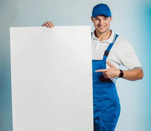 Imagem falsa de um jovem trabalhador sorridente usando uniforme azul. Segurando banner branco vazio em sua mão e apontando o dedo para você olhando para a câmera. Isolado em fundo cinza com espaço de cópia. Expressão facial humana, emoção. Conceito de negócio . — Fotografia de Stock