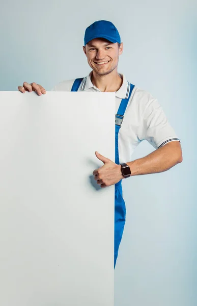 Burla de un joven trabajador sonriente con uniforme azul. Sostenía un estandarte blanco vacío en su mano y mostraba el pulgar hacia arriba. Movimiento fresco. Aislado sobre fondo gris con espacio de copia. Expresión de rostro humano, emoción. Concepto empresarial . — Foto de Stock