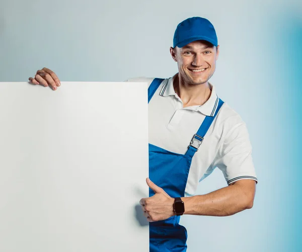 Burla de un joven trabajador sonriente con uniforme azul. Sostenía un estandarte blanco vacío en su mano y mostraba el pulgar hacia arriba. Movimiento fresco. Aislado sobre fondo gris con espacio de copia. Expresión de rostro humano, emoción. Concepto empresarial . — Foto de Stock