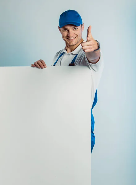 Attrappe Bild eines jungen lächelnden Arbeiters in blauer Uniform. Er hält ein weißes, leeres Banner in der Hand und zeigt mit dem Finger in die Kamera. isoliert auf grauem Hintergrund mit Kopierraum. menschlicher Gesichtsausdruck, Emotionen. Geschäftskonzept. Stockbild