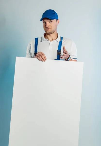 Attrappe Bild eines jungen lächelnden Arbeiters in blauer Uniform. Er hält ein weißes, leeres Banner in der Hand und zeigt mit dem Finger in die Kamera. isoliert auf grauem Hintergrund mit Kopierraum. menschlicher Gesichtsausdruck, Emotionen. Geschäftskonzept. lizenzfreie Stockbilder