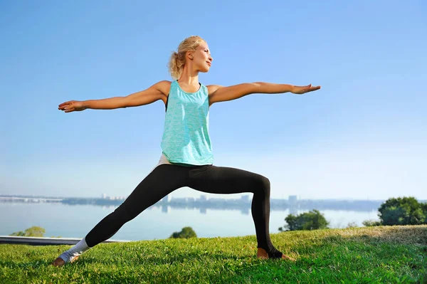 Woman practicing warrior yoga pose at the fresh lawn at the rive