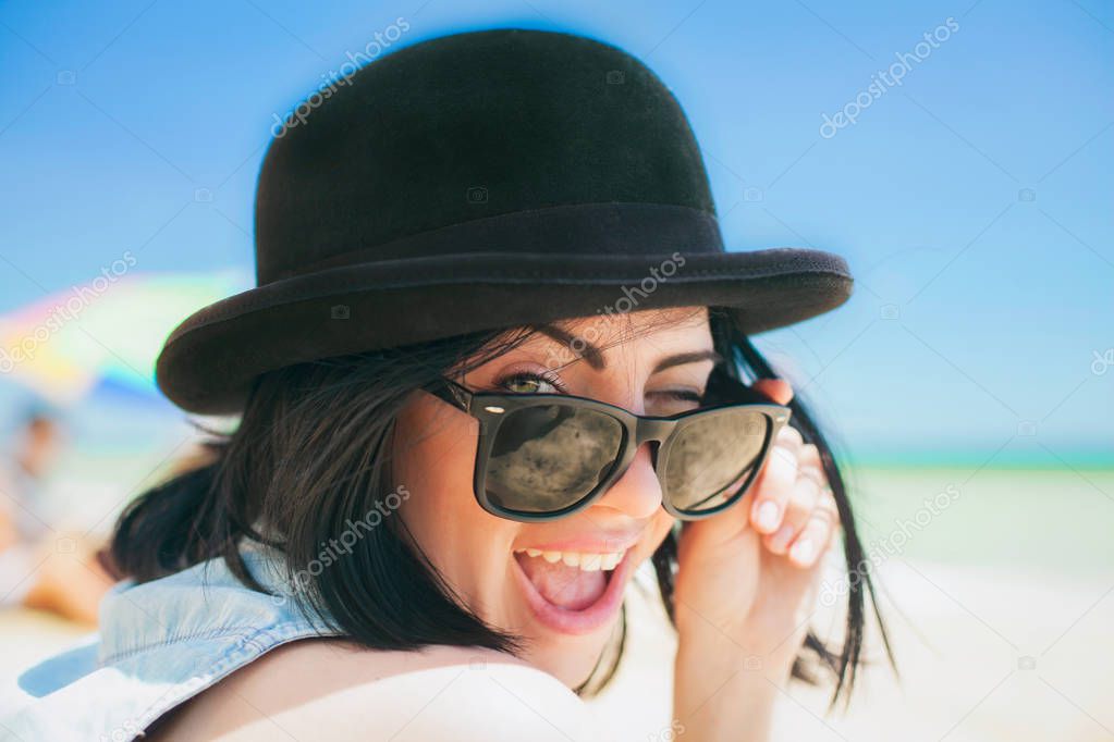 Portrait of smiling beautiful young woman in black hat and retro sunglasses on the beach