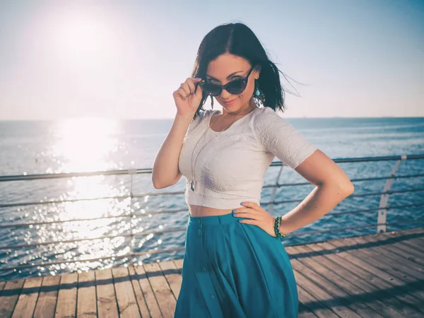 Mooie jonge vrouw die zich voordeed als model permanent aan kade in de buurt van de zee of Oceaan. Lady dragen retro stijl rok en zonnebril. Ochtendzon. — Stockfoto