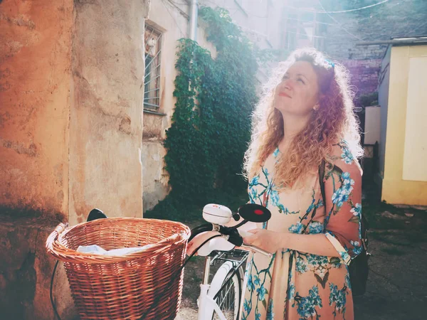 Vacker ung kvinna som bär lång klänning står nära retro vit cykel med korg och tittar på bågen i gamla gård — Stockfoto