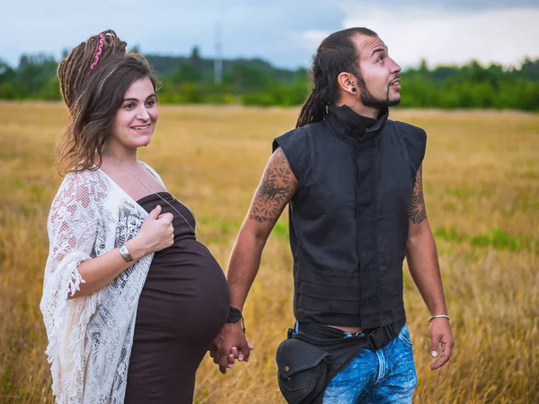 Man in een zwart doek en een zwangere vrouw in lange jurk met dreadlocks worden gehouden in de hand, tegen de achtergrond van blauwe hemel, aard, herfst of zomer seizoen. het verhaal van de liefde. Informele mensen met een tattoo een — Stockfoto