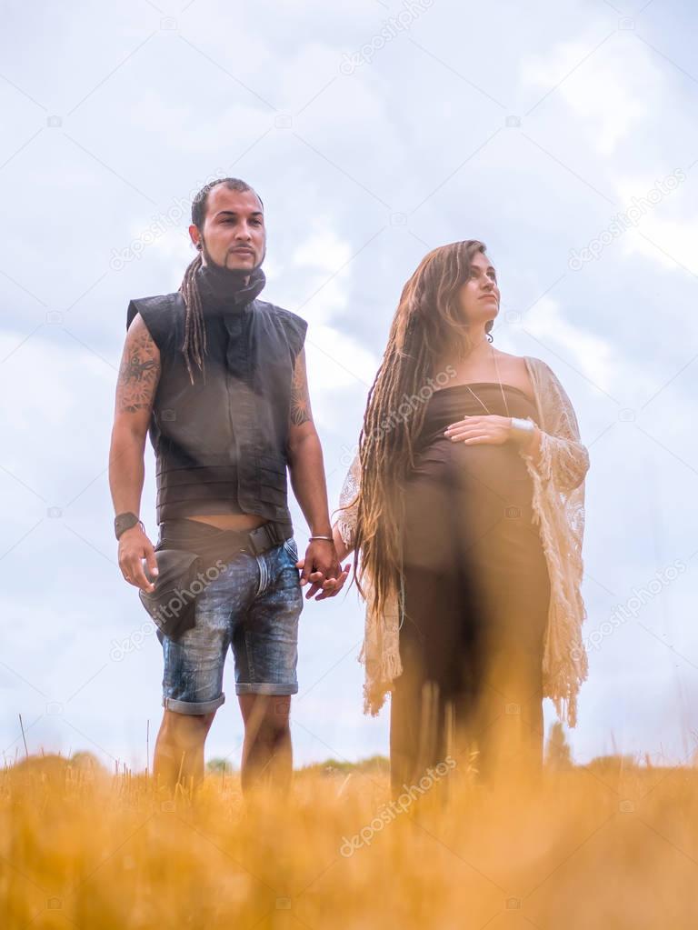 man in a black cloth and a pregnant woman in long dress with dreadlocks are kept in hand, against the background of blue sky, nature, autumn or summer season. love story. Informal people with tattoo a