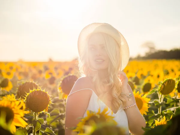 Junge schöne blonde Frau, die im Sonnenblumenfeld steht. Hintergrund Sonnenuntergang. sexy sinnliches Porträt eines Mädchens mit Strohhut und weißem Sommerkleid. — Stockfoto