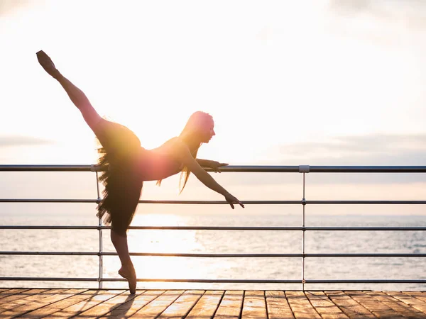 Egy gyönyörű ballerina teljesítő piruett. Szőke nő fekete balett tutu töltésén napkeltekor óceán vagy tenger strand felett. Női tánc karral. — Stock Fotó