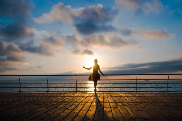 Silueta de bailarina bailadora en tutú de ballet negro y punta en terraplén sobre el océano o el mar al amanecer o al atardecer. Mujer rubia atractiva joven con pelo largo practicando estiramientos y ejercicios . — Foto de Stock