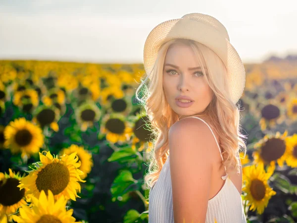 Молода красива блондинка, що стоїть на соняшниковому полі. Тло заходу сонця. Сексуальний чуттєвий портрет дівчини в солом'яному капелюсі та білій літній сукні . — стокове фото