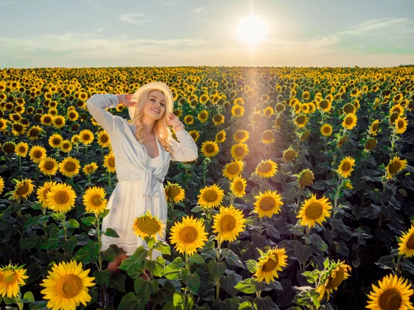 Młody piękny blond kobiety stojącej w Słonecznikowe pole. Zachód słońca na tle. Portret zmysłowy sexy dziewczyna w słomkowym kapeluszu i biały Letnia sukienka. — Zdjęcie stockowe