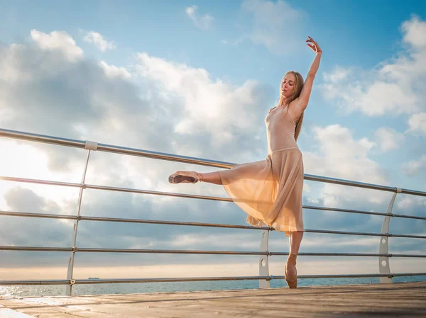 Ballerina danzante in abito di seta beige e punta sul terrapieno sopra l'oceano o la spiaggia al sorgere del sole. Giovane bella donna bionda con i capelli lunghi praticare esercizi classici con emozioni . — Foto Stock