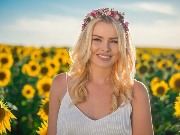 Junge schöne blonde Frau, die im Sonnenblumenfeld steht. Hintergrund Sonnenuntergang. sexy sinnliches Porträt eines Mädchens mit Blumenkranz und weißem Sommerkleid. — Stockfoto