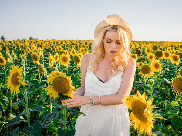 Junge schöne blonde Frau, die im Sonnenblumenfeld steht. Hintergrund Sonnenuntergang. sexy sinnliches Porträt eines Mädchens mit Strohhut und weißem Sommerkleid. — Stockfoto