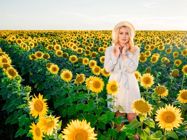 Tânără frumoasă blondă în picioare în câmp de floarea-soarelui. Apus de soare. Sexy senzual portret de fată în pălărie de paie și rochie de vară albă . — Fotografie, imagine de stoc