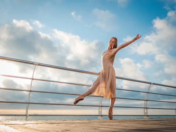 Bailarina dançante em vestido de seda bege e ponteiro no dique acima do oceano ou praia do mar ao nascer do sol. Jovem mulher loira bonita com cabelos longos praticando exercícios clássicos com emoções . — Fotografia de Stock