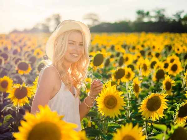 Giovane bella donna bionda in piedi nel campo di girasole. Sfondo del tramonto. Sexy ritratto sensuale di ragazza in cappello di paglia e bianco abito estivo . — Foto Stock