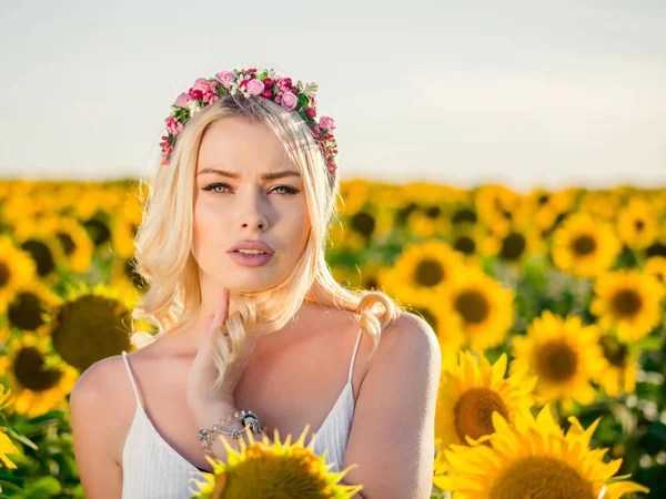 Jovem mulher loira bonita de pé no campo de girassol. Pôr-do-sol. Sexy retrato sensual da menina em flores grinalda e vestido de verão branco . — Fotografia de Stock