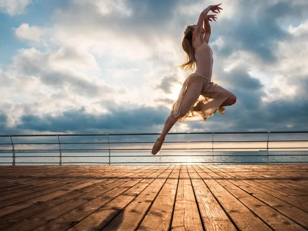 Jumping ballerina in beige dress and pointe on embankment above ocean or sea beach at sunrise.Beautiful blonde woman with long hair practicing stretching and classic exercises.Epic jump.