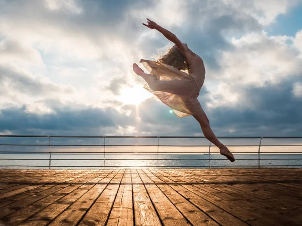 Salto ballerina in abito beige e punta su argine sopra l'oceano o spiaggia di mare al sunrise.Beautiful donna bionda con i capelli lunghi che praticano stretching e esercizi classici.Salto epico . — Foto Stock